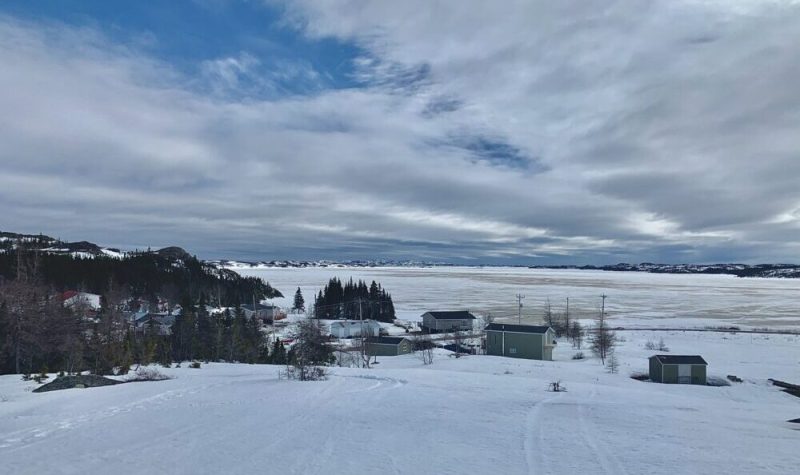 Sous un ciel bleu et blanc, quelques maisons aux abords d'une baie encore enneigée.