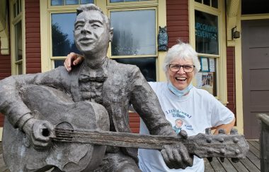 A smiling woman has her arm around a statue of a man playing guitar