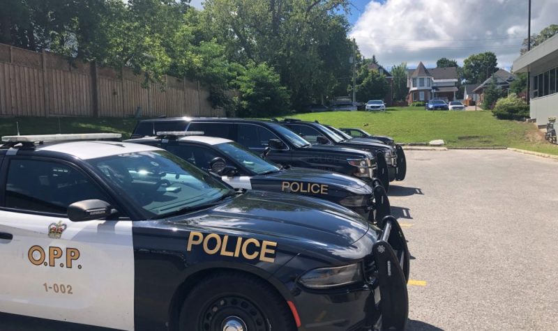 On peut voir une série de véhicules de police de la Police provinciale de l'Ontario. Les voitures sont blanches et noires avec les inscriptions de la police sur le flanc. À l'arrière-plan, on peut voir le stationnement du poste de police de Midland.