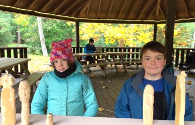 Two children sit behind a table with wooden carvings in front of them.