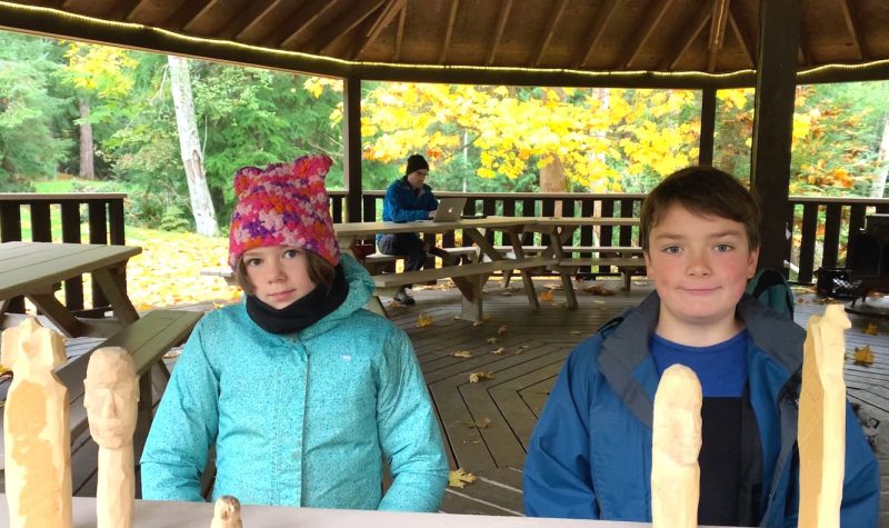 Two children sit behind a table with wooden carvings in front of them.