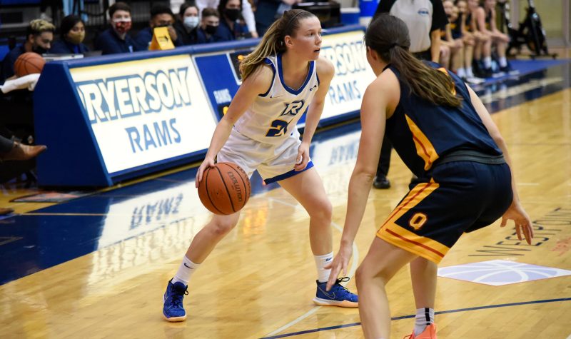 A woman in a white and blue basketball uniform dribbles a ball on a court during a game.