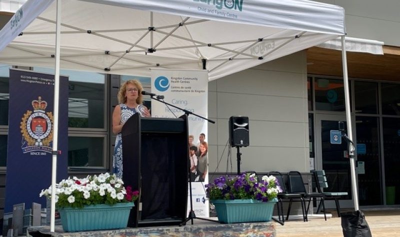 A woman speaks into a microphone under a tent, with the Kingston Police logo and the Kingston Community Health Centres banners behind her.