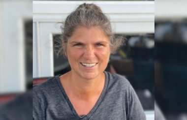 A woman with salt-and-pepper hair and a heather grey V-neck shirt smiles at the camera in a portrait shot that appears to have been taken indoors.