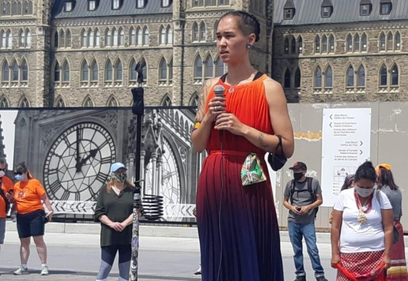 NDP MP Mumilaaq Qaqqaq stands before Parliament addressing a crowd with a microphone in hand.