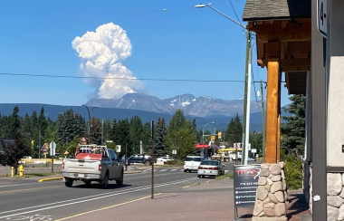 Cars and trucks travel on a small town street as a fire plume over a mountain range can be seen in the near distance.
