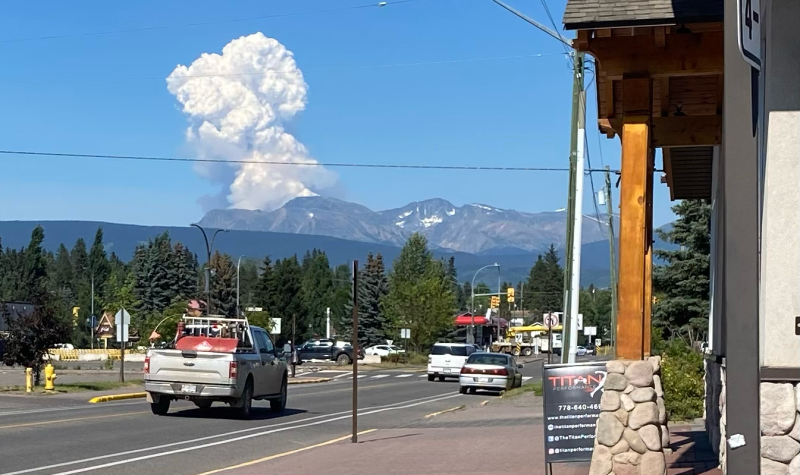 Cars and trucks travel on a small town street as a fire plume over a mountain range can be seen in the near distance.