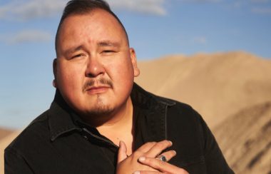 A professional head shot of singer songwriter William Prince. In the background, there is a blue sky and sandy mountain.