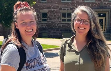 Two women are shown smiling as they stand in the sunshine outside a university building.