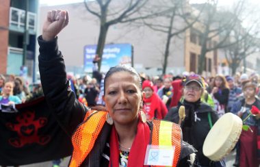 Cintesapa Taweya (aka Juanita Desjarlais) stands with her fist in the air with people marching behind her and playing drums