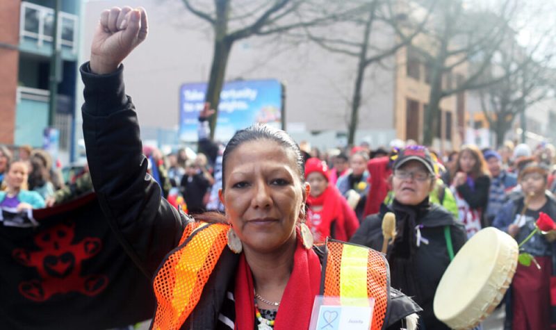 Cintesapa Taweya (aka Juanita Desjarlais) stands with her fist in the air with people marching behind her and playing drums