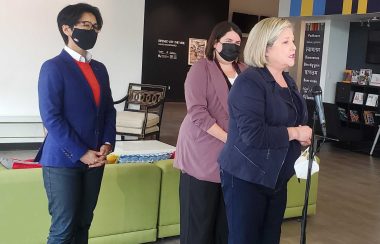 Three people standing in front of a green couch as they speak to a microphone. One person has a blue blazer, the person in the middle has a lavender coat and the person on the far right wears a navy jackey and has blonde hair.