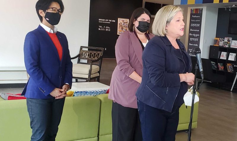 Three people standing in front of a green couch as they speak to a microphone. One person has a blue blazer, the person in the middle has a lavender coat and the person on the far right wears a navy jackey and has blonde hair.