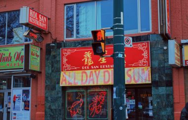 A yellow and red sign on top of a storefront with various other signs around it. A stop light is shown in front of the red and yellow sign.