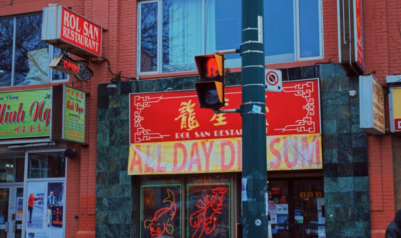 A yellow and red sign on top of a storefront with various other signs around it. A stop light is shown in front of the red and yellow sign.