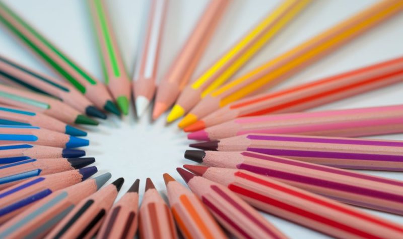 On peut voir une série de crayon de couleur en bois placés en cercle sur un fond blanc. L'ensemble donne un peu l'effet du drapeau multicolore de la fierté