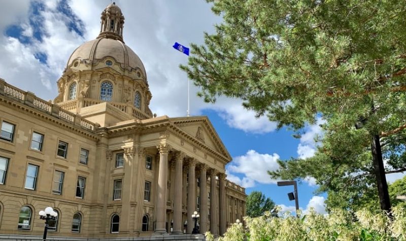 The Alberta Legislature from the front, a tree sits in the photo too. The Alberta flag waves on top of the building. Weather is partly cloudy.