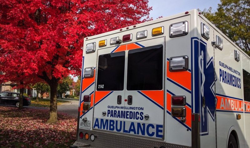 An ambulance sits by a curb with fall leaves and a red-leaved tree in the background.