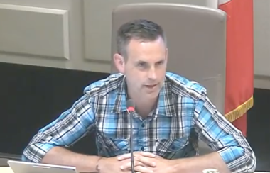 Andrew Black sits at a white desk during a Sackville town council meeting with a Canadian flag behind him and a lap top and microphone in front of him