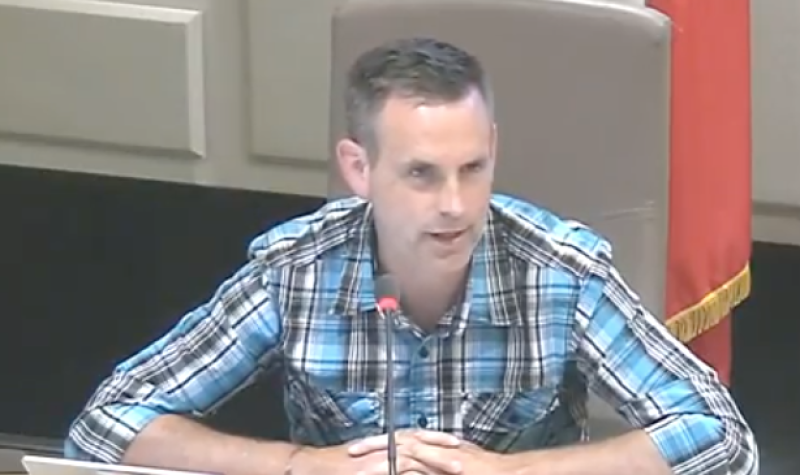 Andrew Black sits at a white desk during a Sackville town council meeting with a Canadian flag behind him and a lap top and microphone in front of him