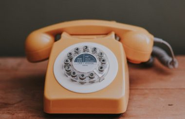 On voit un téléphone traditionnel orange de style « téléphone à roulette » des années 1980. Le téléphone est posé sur une table en bois.