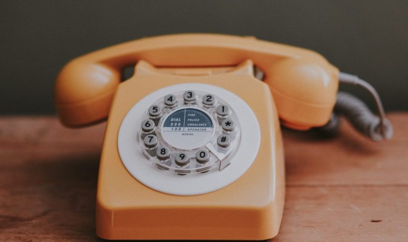 On voit un téléphone traditionnel orange de style « téléphone à roulette » des années 1980. Le téléphone est posé sur une table en bois.