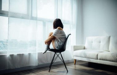 Une jeune femme, accroupie sur une chaise blanche, regarde par la fenêtre.