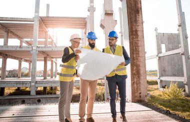 Three men stand looking at a construction blueprint. The men all are wearing yellow reflective vests as well as hard hats. The men are standing in a construction zone with a partly constructed building in the background.