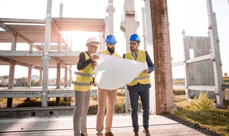 Three men stand looking at a construction blueprint. The men all are wearing yellow reflective vests as well as hard hats. The men are standing in a construction zone with a partly constructed building in the background.