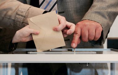 Hands putting envelop into ballot box while a mans finger presses button to count the ballot.