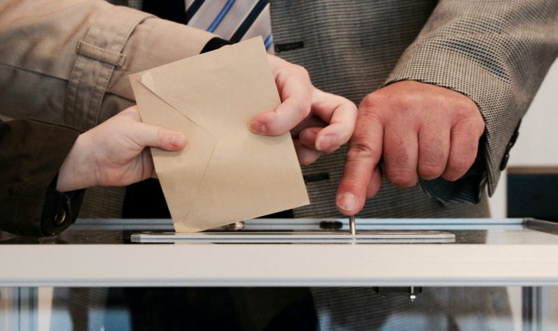 Hands putting envelop into ballot box while a mans finger presses button to count the ballot.