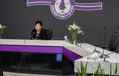 A woman with short black hair sits behind a white and purple table. On the grey wall behind the table is a purple logo which says 'Six Nations Grand River'