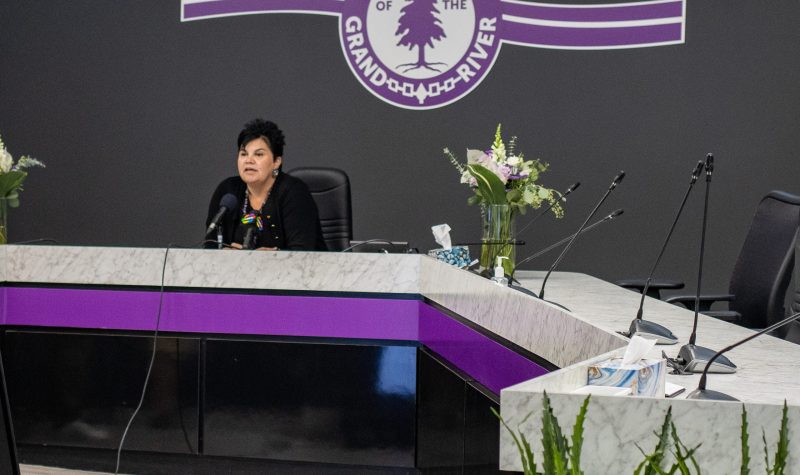 A woman with short black hair sits behind a white and purple table. On the grey wall behind the table is a purple logo which says 'Six Nations Grand River'