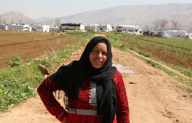 A person in a red dress and black burka. They are standing on a dirt path in a field.