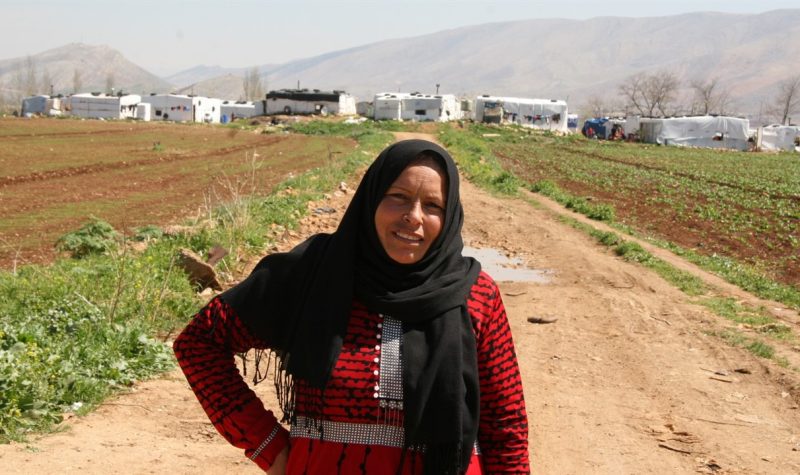 A person in a red dress and black burka. They are standing on a dirt path in a field.