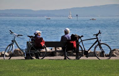 Deux personnes âgées sur un banc face à la mer, des vélos sont posé sur le banc, ils ont l'air de faire une pause. Ils ont l'air actifs.