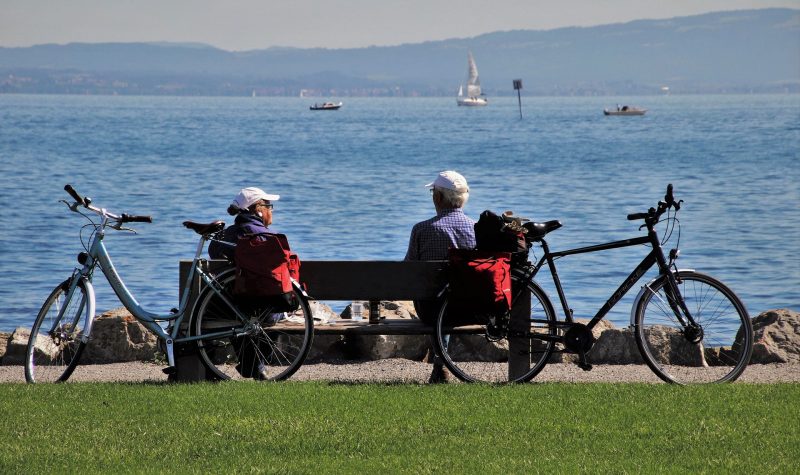 Deux personnes âgées sur un banc face à la mer, des vélos sont posé sur le banc, ils ont l'air de faire une pause. Ils ont l'air actifs.