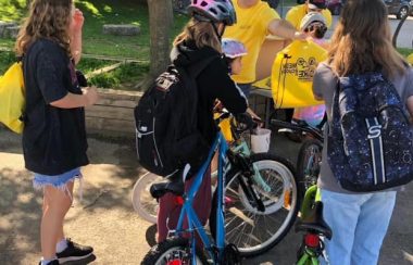 Volunteers is yellow t shirts hand out yellow prize bags to students on bikes in a school parking lot