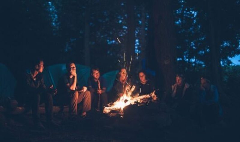 Dans la pénombre, de nombreux jeunes sont assis autour d'un feu de camp au coeur d'une forêt.