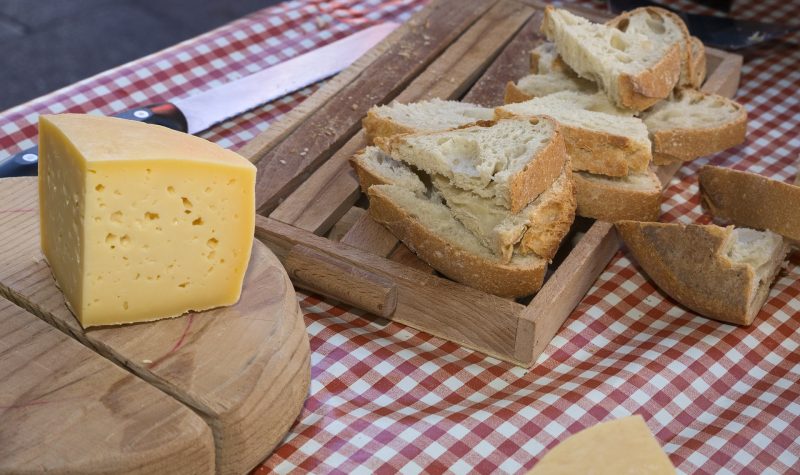 A block of cheese sits on a wooden plate. A loaf of bread sits on another wooden plate. The plates are sitting on a red and white checkered table.