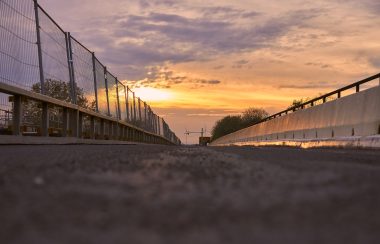 A view of roadway on bridge. an asphalt barrier sits on the right side of the road and a fenced barrier sits on the left. The sun sets in the background.