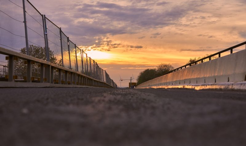 A view of roadway on bridge. an asphalt barrier sits on the right side of the road and a fenced barrier sits on the left. The sun sets in the background.