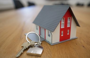 Petite maison miniature de couleur blanche et rouge avec un toit gris, avec un porte-clé avec une seule clé à côté, déposés sur une table.