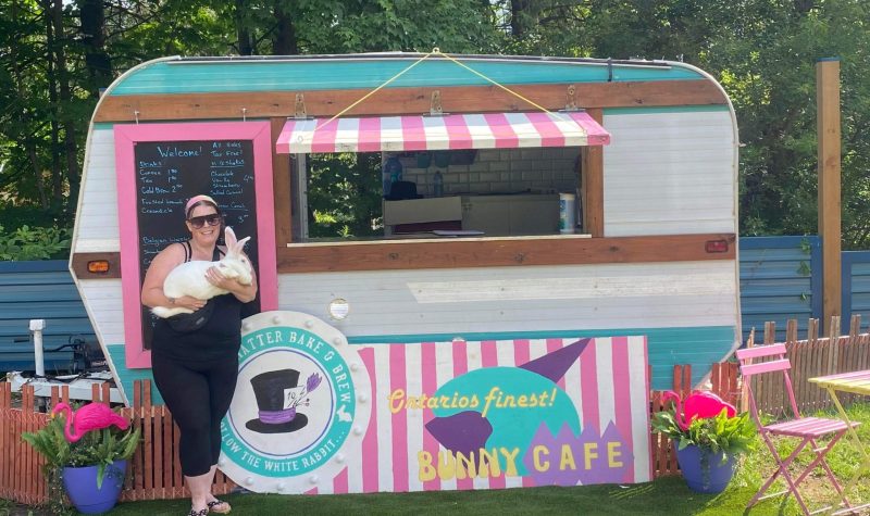 A bright pink and teal food truck sits in a vibrant outdoor area. A woman holds a large white rabbit in front of it.