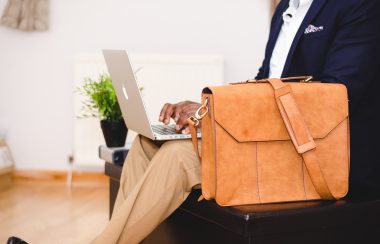 Une personne assise dans un fauteuil avec un ordinateur sur ses genoux et un sac de voyage devant. La personne est bien habillée et on ne voit pas son visage. l'image est très lumineuse.