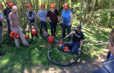 A group of people with hard hats are gathered outside around a man demonstrating an orange water pump with a long black hose.