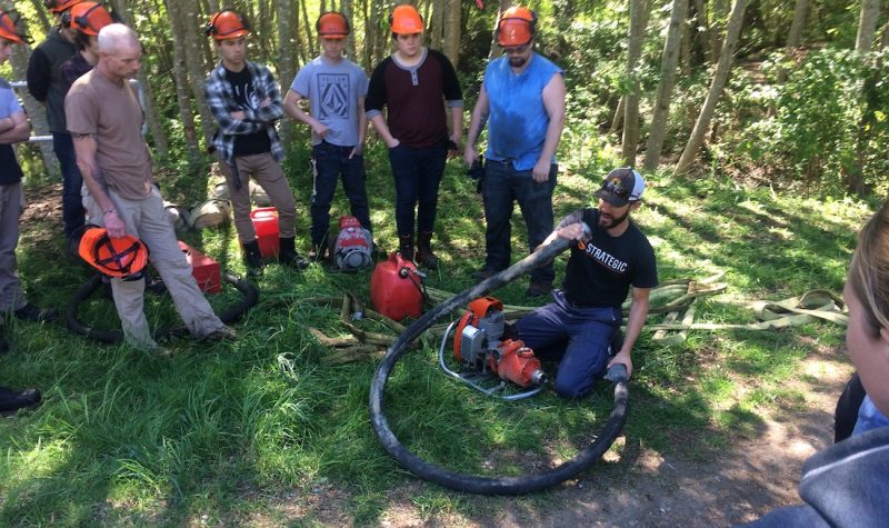 A group of people with hard hats are gathered outside around a man demonstrating an orange water pump with a long black hose.