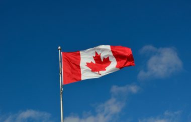 A Canadian flag blowing in the wind. Blue skies surround the flag.