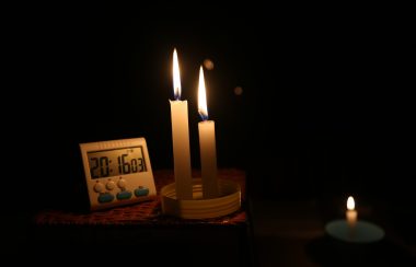 Two long candles sit on a table lit. beside them sits a electronic 24 hour clock. A smaller candles sits to the right of the 2 other candles.
