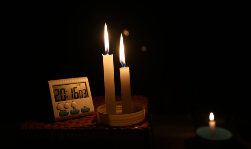 Two long candles sit on a table lit. beside them sits a electronic 24 hour clock. A smaller candles sits to the right of the 2 other candles.